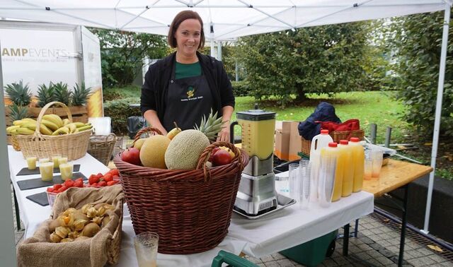 Eine junge Frau steht am Smoothie Stand vor einer reich gedeckten Fläche von Obst und Gemüse.