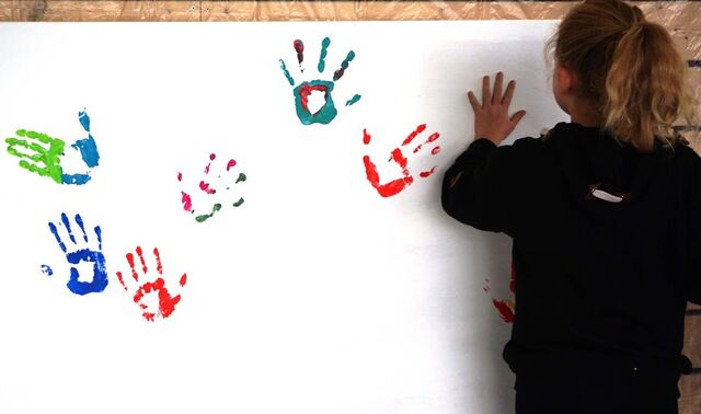 Ein großes, dunkelhaariges Mädchen ist von hinten zu sehen, wie sie ihren Handabdruck auf eine große Leinwand mit bunten Handabdrücken macht.