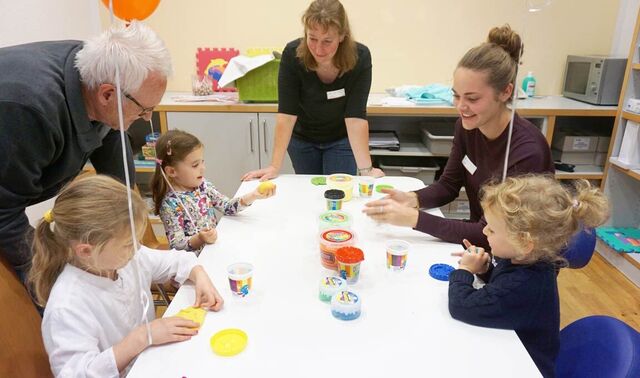 Zwei junge Therapeutinnen sitzen mit einer Gruppe kleiner Kinder am Tisch. Die Kinder kneten. Ein äletere Mann beugt sich zu einem der Kinder.