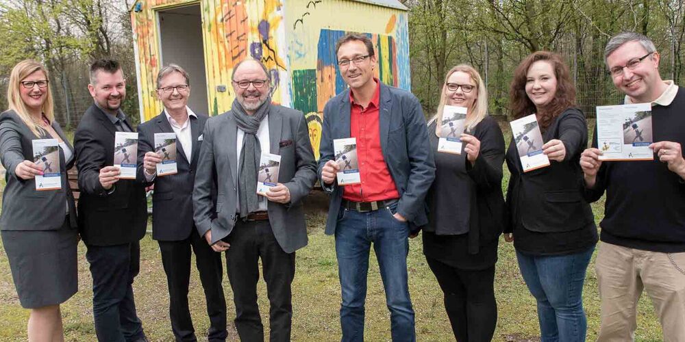 Team schaut in die lächelnd in die Kamera und jeder hält einen Flyer der Augustinus Gruppe in der Hand. und zeigt diesen in die Kamera.